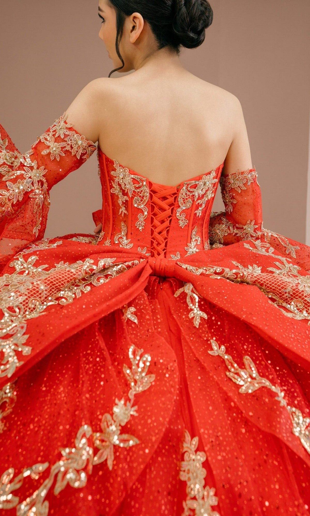 A woman is seen from behind wearing the Quinceanera Dress 8575JQ by Cinderella Coutureโa luxurious red and gold embellished ball gown featuring a lace-up back and intricate floral embroideryโset against a neutral background.